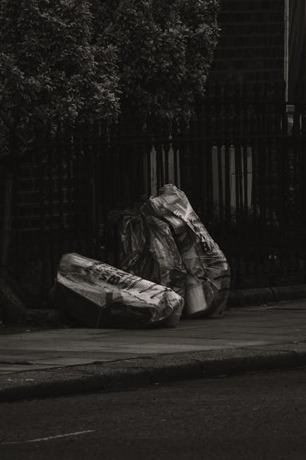 In the foreground, three large black plastic rubbish bags, filled and loosely tied, rest on a street pavement near a curb, with some of the bags exhibiting creases and reflective surfaces that catch m