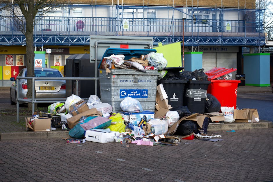 A small blue flatbed truck parked on the side of a road, heavily loaded with a variety of discarded rubbish materials, including large cardboard boxes, plastic bags, and miscellaneous debris. The truc